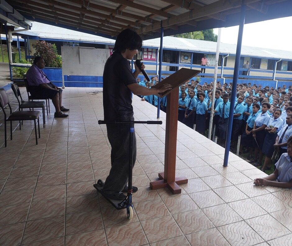 学校の全校集会で、スクーターを手に生徒たちへ講演する様子